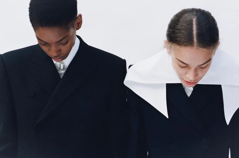 Two young fashion models in black blazers, standing side-by-side against a white background, showcasing a minimalist and modern aesthetic.
