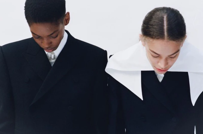 Two young fashion models in black blazers, standing side-by-side against a white background, showcasing a minimalist and modern aesthetic.