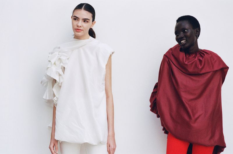 Two fashion models pose in front of a white backdrop; one wears a white top with ruffles, and the other wears a maroon top with red pants.