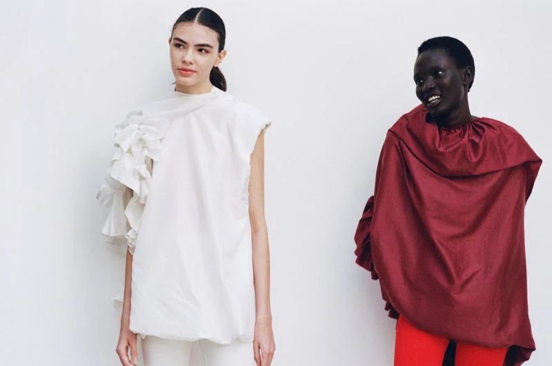 Two fashion models pose in front of a white backdrop; one wears a white top with ruffles, and the other wears a maroon top with red pants.