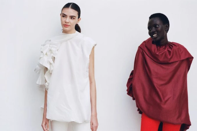 Two fashion models pose in front of a white backdrop; one wears a white top with ruffles, and the other wears a maroon top with red pants.