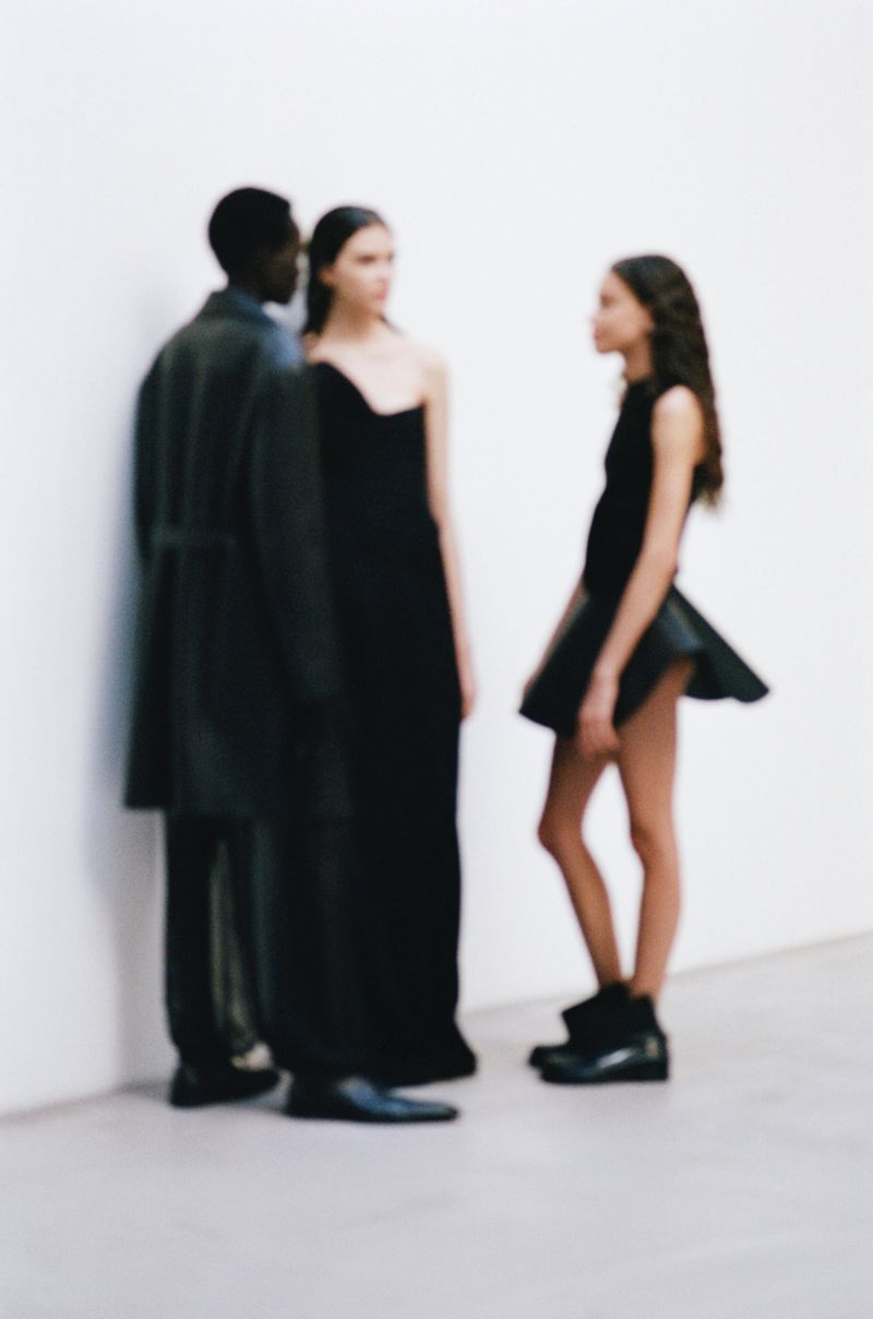 Three fashion models in dark gowns and coat pose against a neutral background in a studio fashion shot for a minimalist concept.