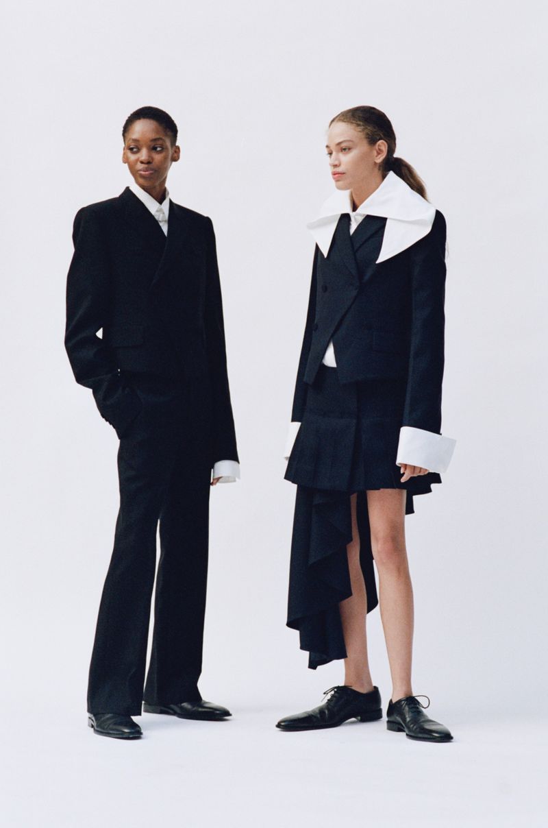 Two women in formal attire pose in a photo studio. The one on the left wears a pantsuit, while the other is in a skirt suit.