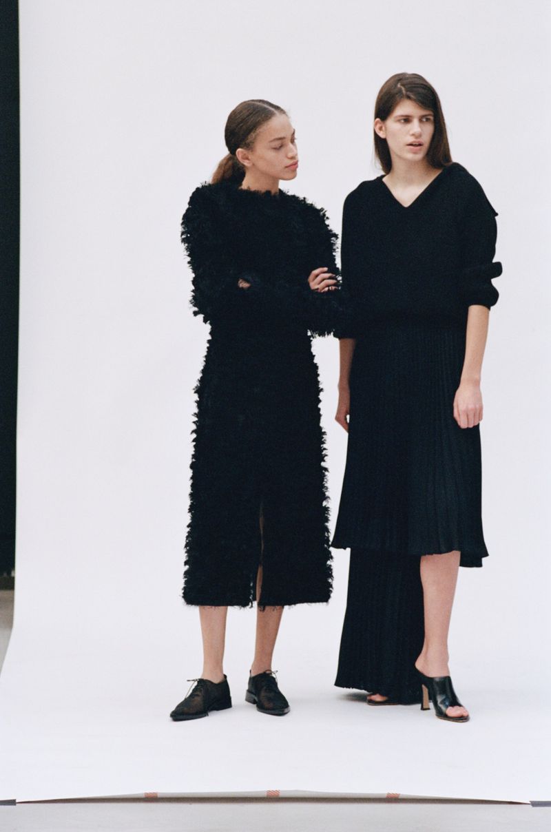 Two fashion models in black dresses stand in a bright studio setting. One model has her arms crossed, while the other stands with a straight posture.