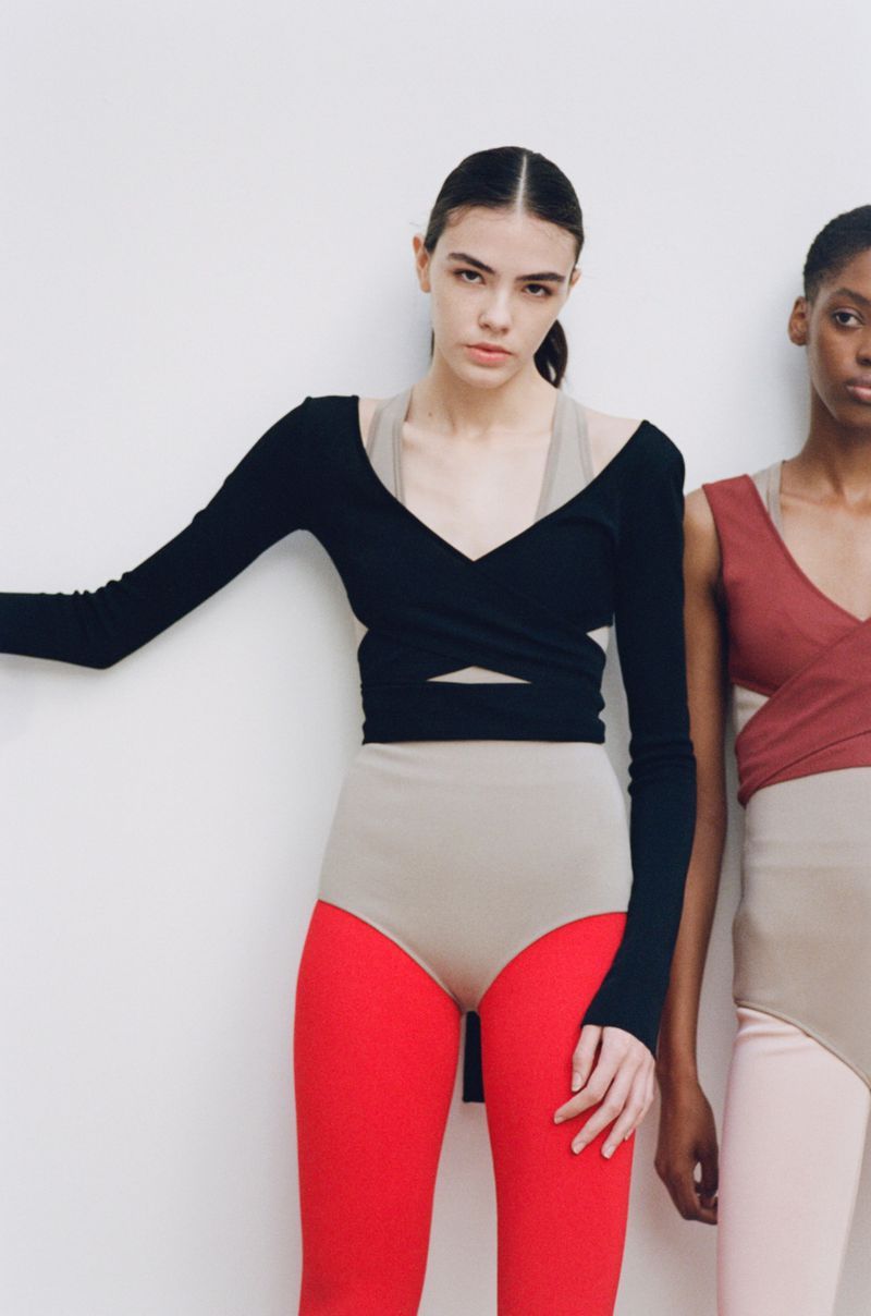 Two women pose in fashionable athletic-wear, featuring a long-sleeve black wrap top, a nude leotard, and bright red leggings against a white backdrop.