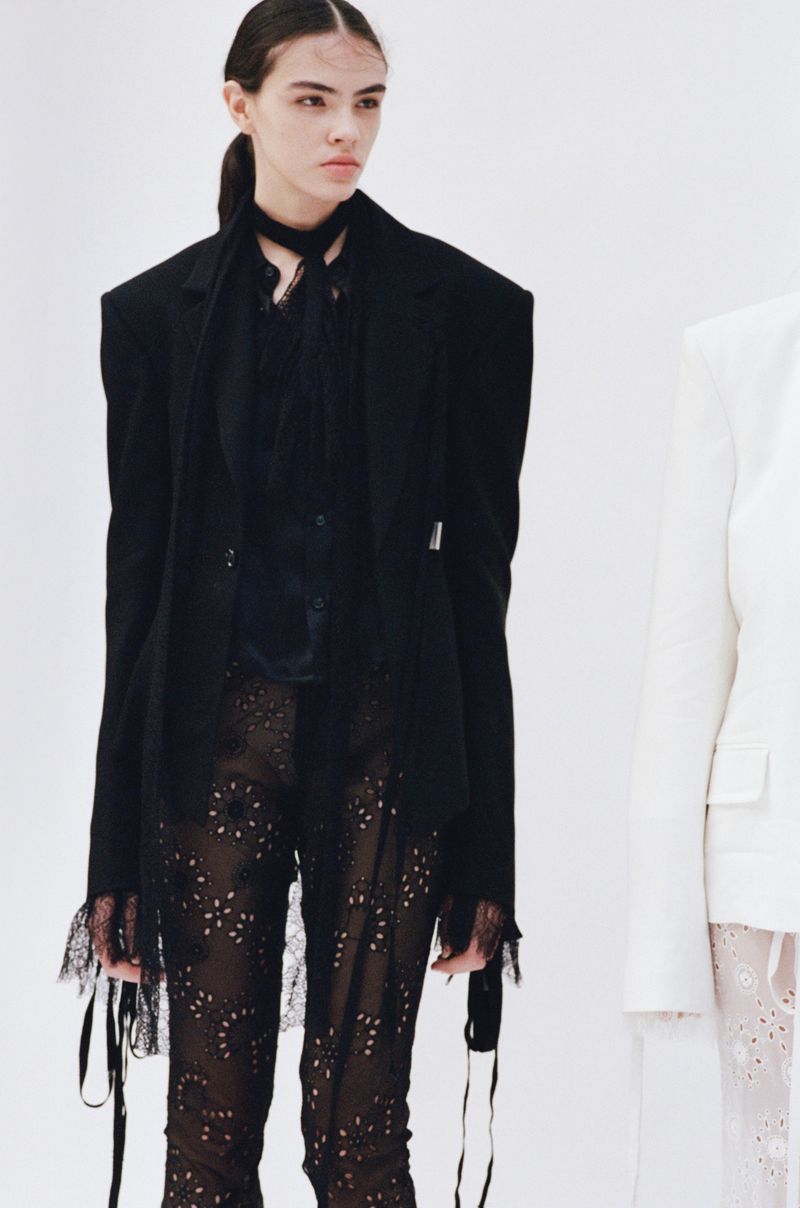 A fashion shot of a young adult female model in a black blazer and pants, standing in a bright white studio environment, side lit.