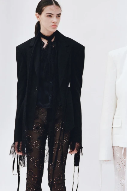 A fashion shot of a young adult female model in a black blazer and pants, standing in a bright white studio environment, side lit.