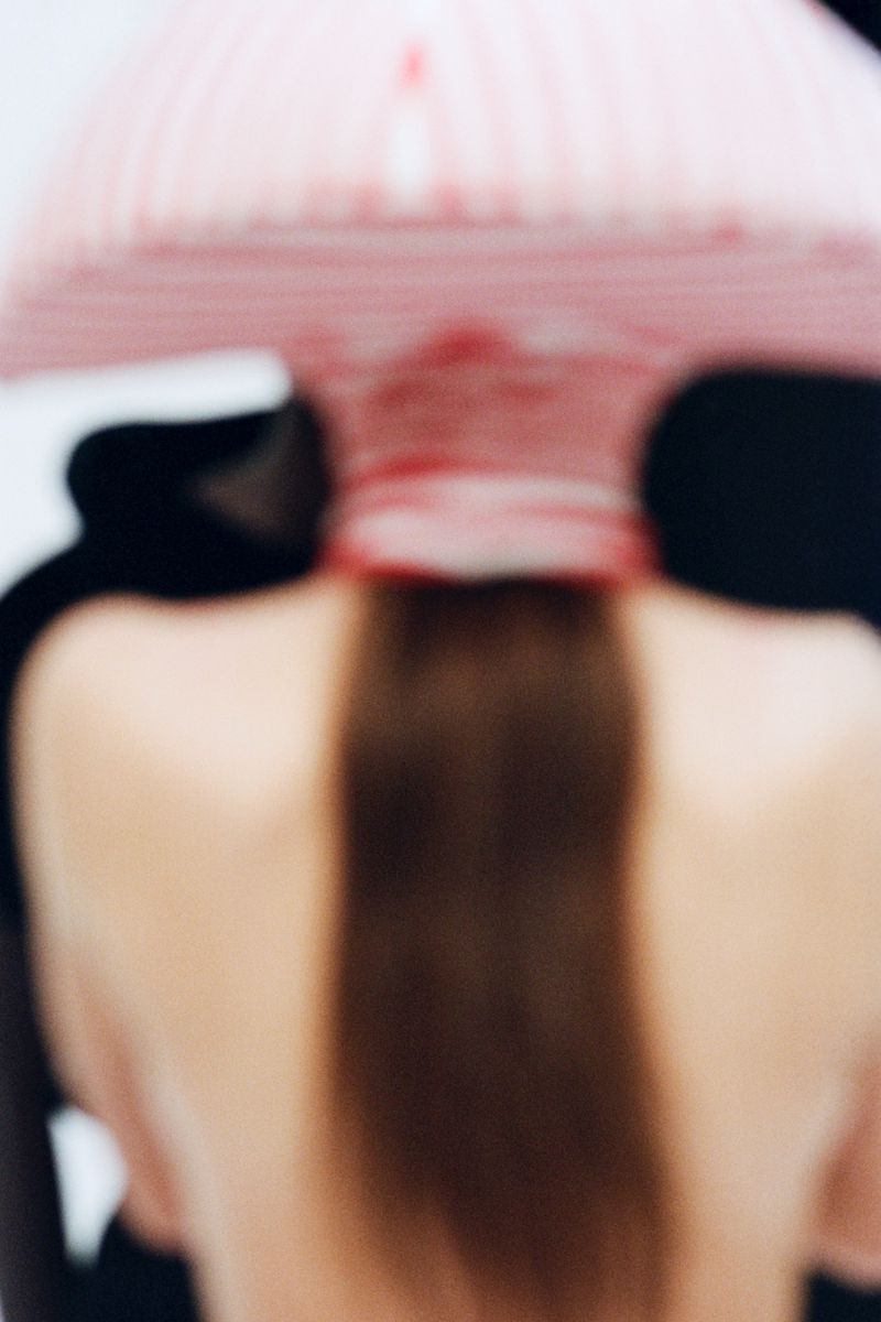 Back view of fair-skinned person with long brown hair wearing striped hat against a white background and a dark shape.