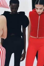 Two female models posing against a white studio background, wearing a black structured top and a red zippered vest.