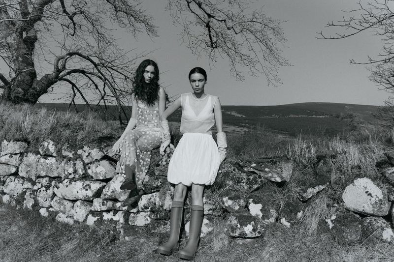 Two women in dresses sit on a stone wall in a rural, black-and-white fashion photo. One wears boots; trees and a hill fill the background.