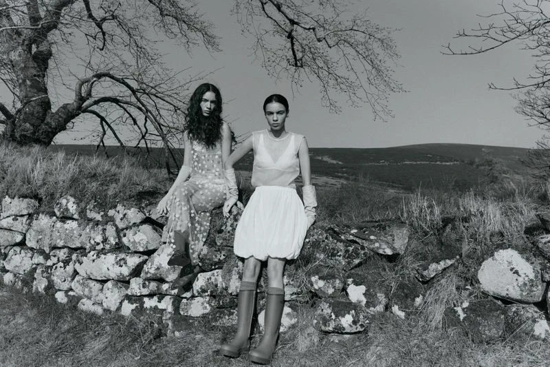 Two women in dresses sit on a stone wall in a rural, black-and-white fashion photo. One wears boots; trees and a hill fill the background.