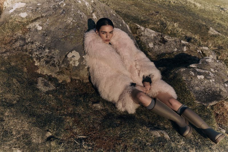 A woman in a pink coat reclines on a rock in an outdoor setting wearing olive green boots with tall socks and minimal makeup.