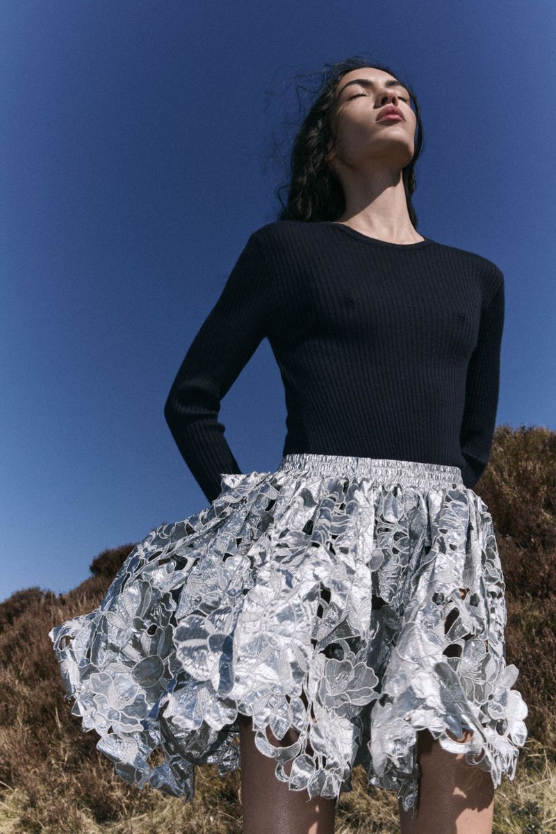An adult female stands outdoors against a blue sky, wearing a long-sleeved black shirt and a silver floral lace skirt in a fashion pose.