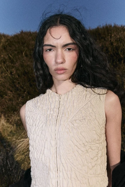 A woman poses outdoors in a textured top against a backdrop of a hill under a clear sky in a medium shot fashion photo.