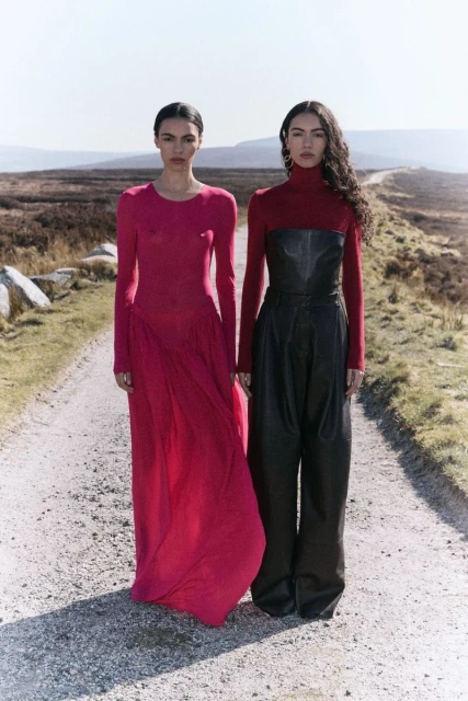 Two women pose in long-sleeve tops and flowing pants on a gravel road. A muted landscape stretches into the distance under a bright sky.