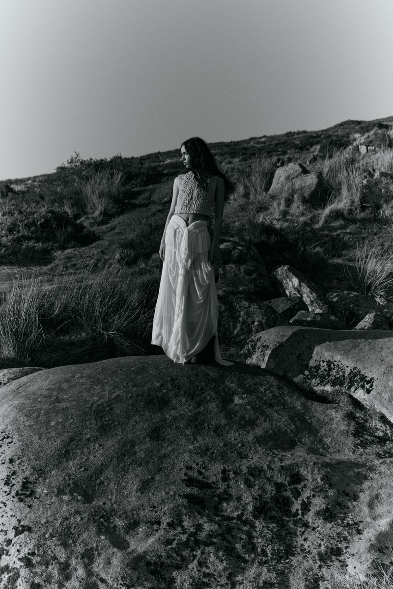 Monochrome photograph of a woman in a flowing white dress standing atop a large rock, set against a grassy hillside and clear sky.