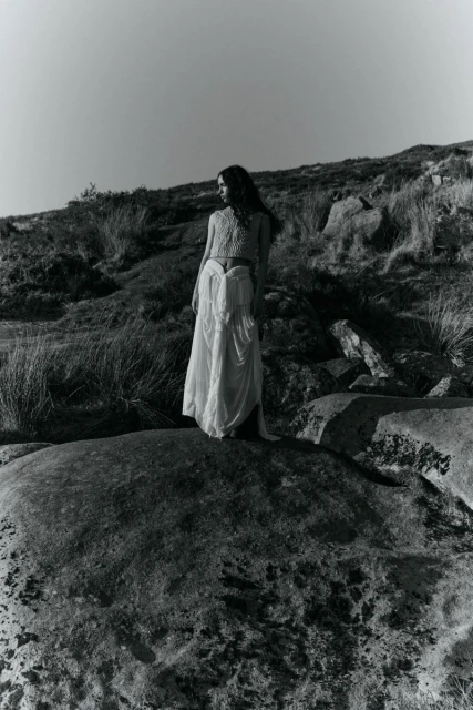 Monochrome photograph of a woman in a flowing white dress standing atop a large rock, set against a grassy hillside and clear sky.