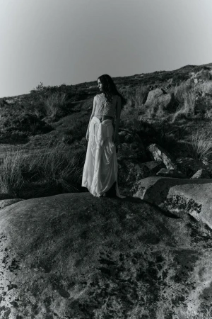Monochrome photograph of a woman in a flowing white dress standing atop a large rock, set against a grassy hillside and clear sky.