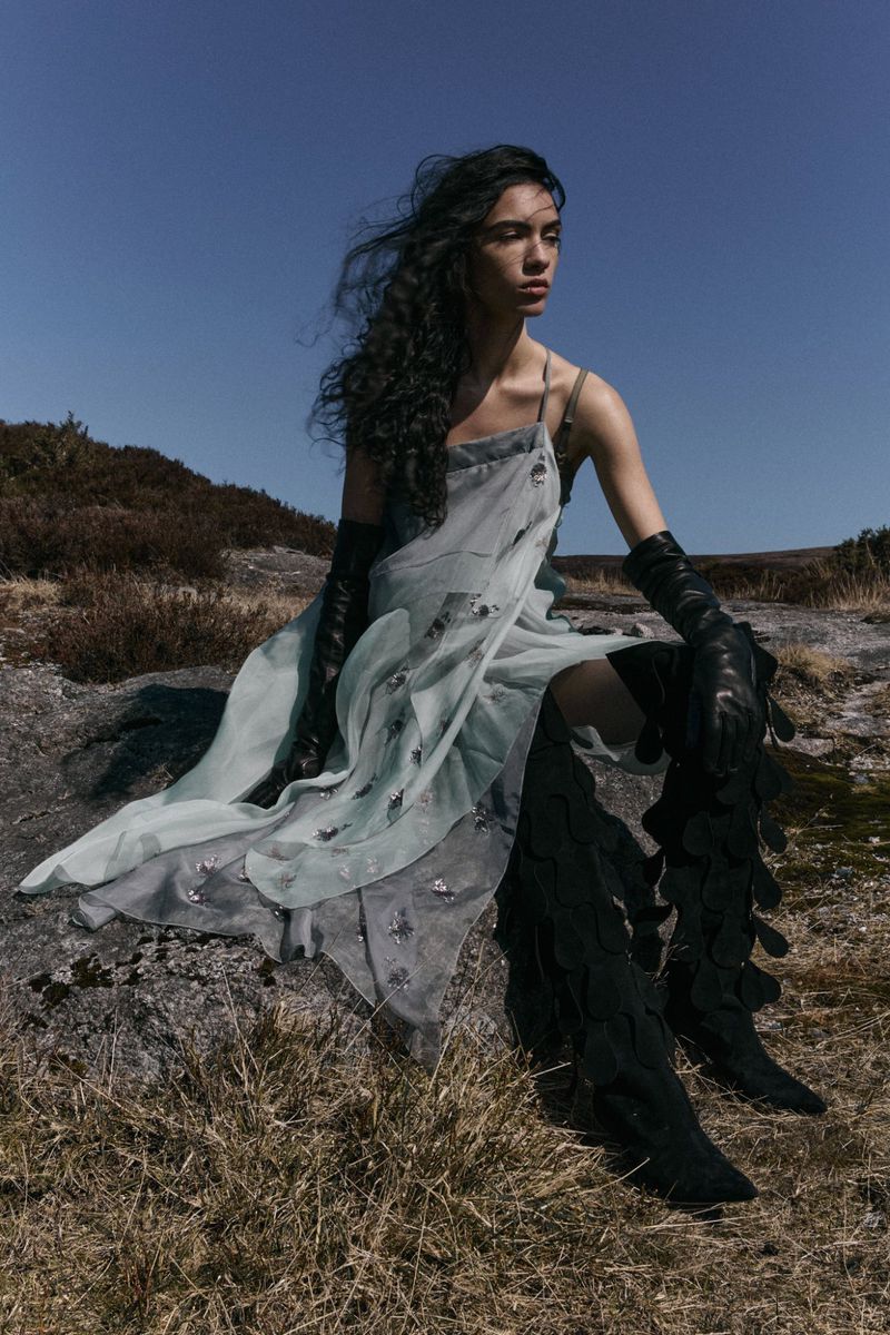 A woman with dark wavy hair poses outdoors, wearing a sheer dress, long gloves, and high boots, seated on a rock in a grassy field.