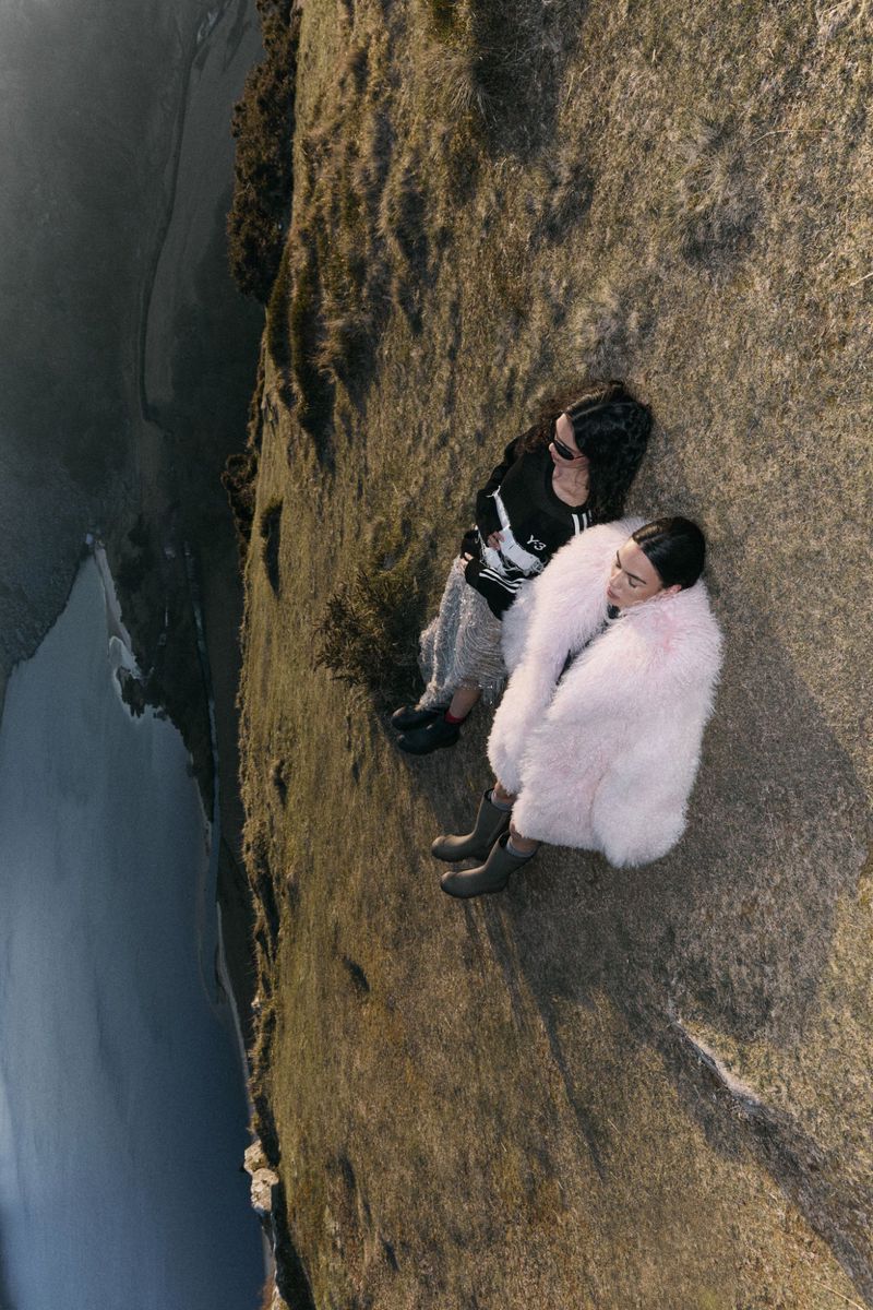 Two women in fashionable outerwear sit on a grassy clifftop overlooking the ocean in a sunlit, high-angle outdoor fashion shot.