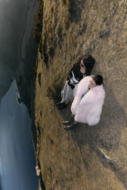 Two women in fashionable outerwear sit on a grassy clifftop overlooking the ocean in a sunlit, high-angle outdoor fashion shot.