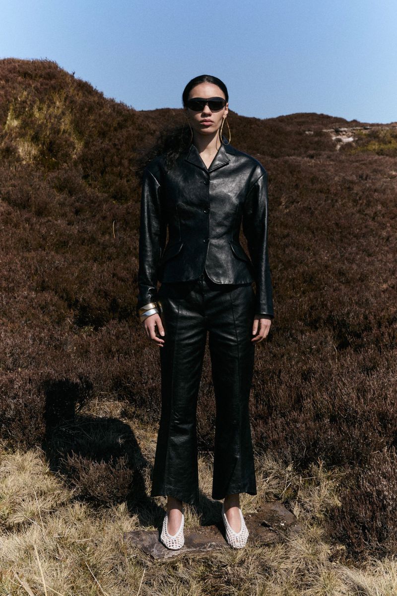 A woman wearing sunglasses and a black leather jacket and pants stands in a field with grassy hills under a clear blue sky.