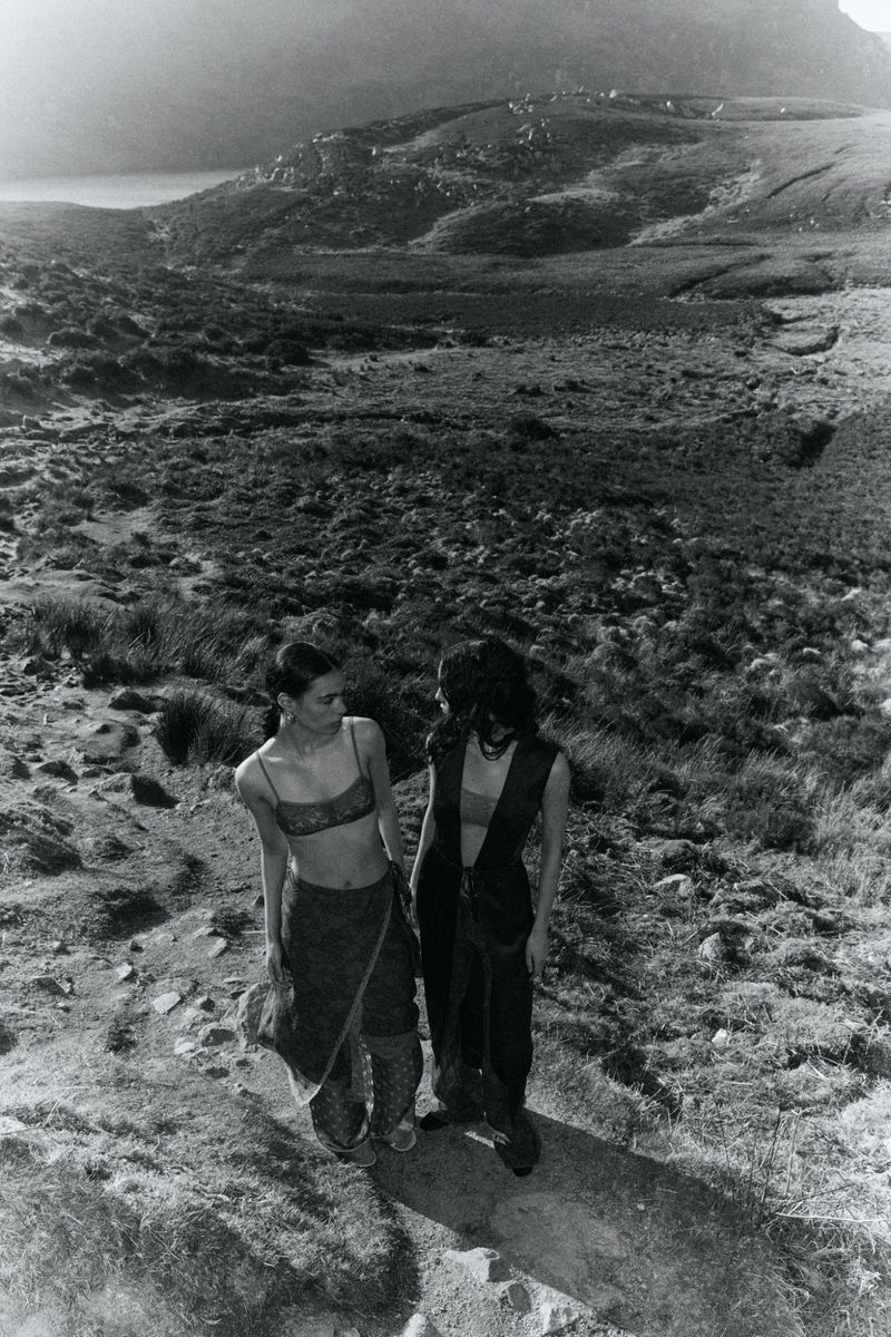 Two women stand on a grassy hill overlooking a valley and mountain range in a black-and-white outdoor fashion editorial shot.