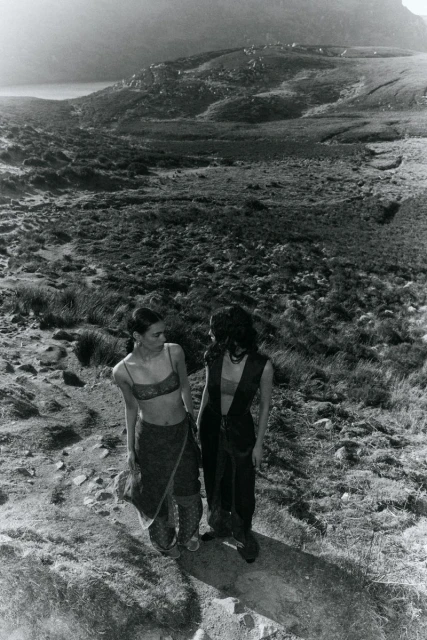 Two women stand on a grassy hill overlooking a valley and mountain range in a black-and-white outdoor fashion editorial shot.