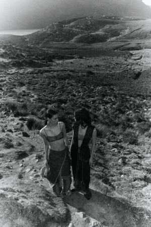 Two women stand on a grassy hill overlooking a valley and mountain range in a black-and-white outdoor fashion editorial shot.