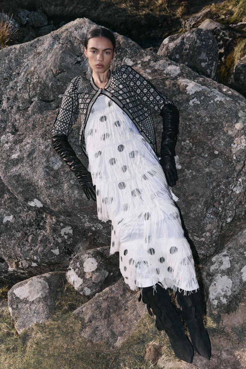Fashion editorial shot of a woman in a fringed dress, jacket, and boots, posing on rocks in a grassy outdoor environment. 