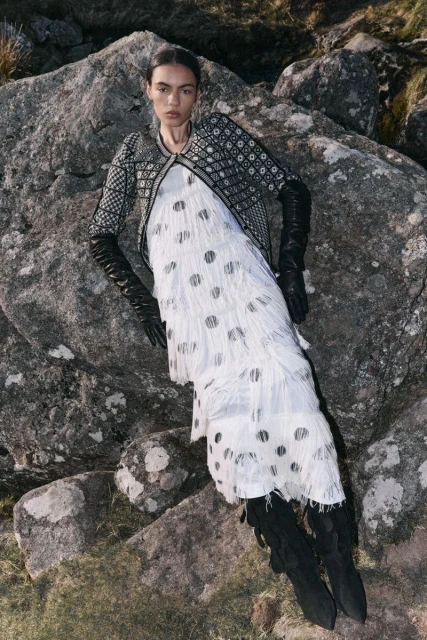 Fashion editorial shot of a woman in a fringed dress, jacket, and boots, posing on rocks in a grassy outdoor environment. 