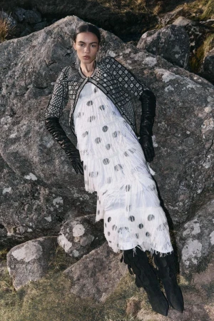 Fashion editorial shot of a woman in a fringed dress, jacket, and boots, posing on rocks in a grassy outdoor environment. 