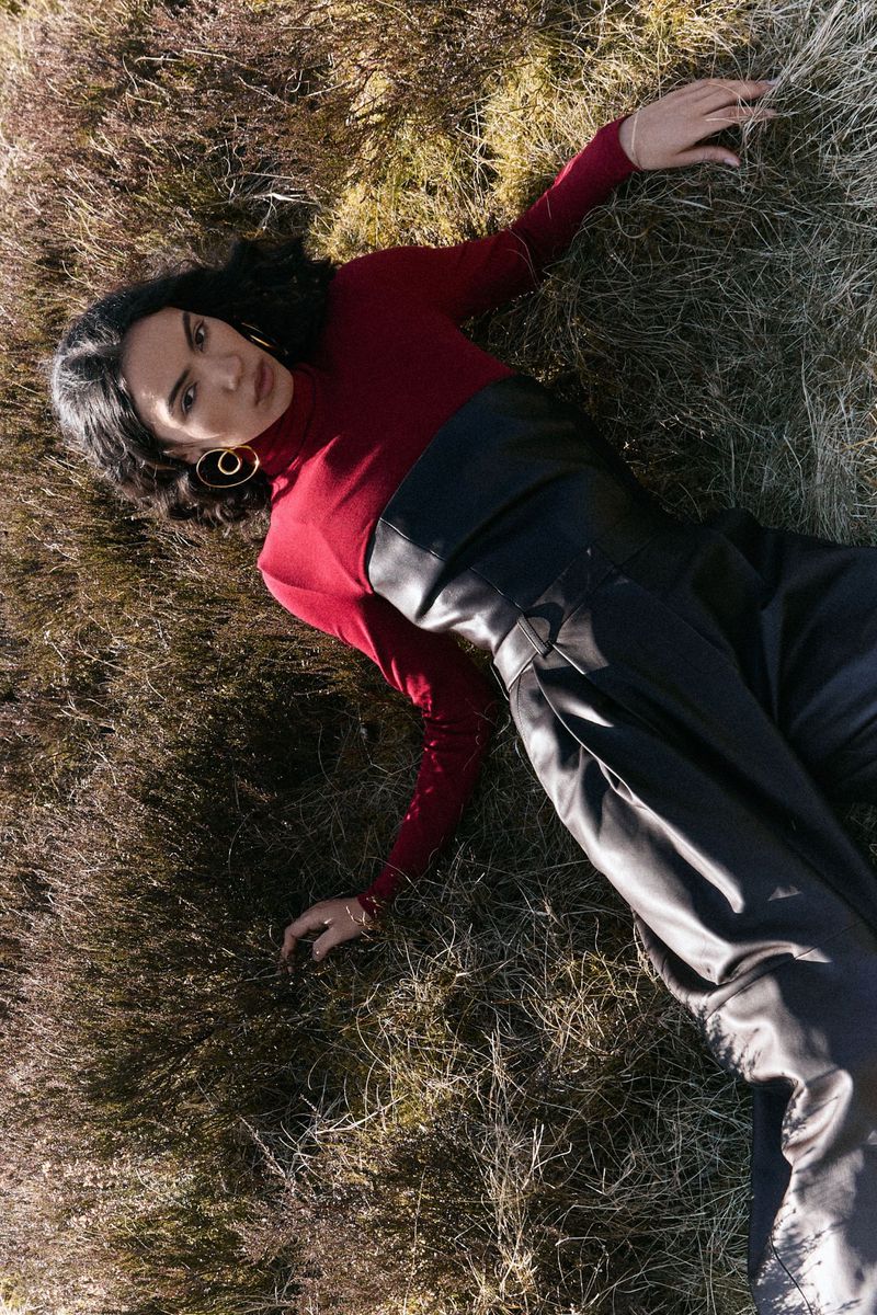Adult female model in a red turtleneck and black pants poses in tall grass. She wears gold hoop earrings and a bracelet.