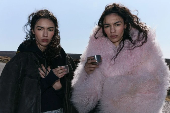 Two women stand outdoors, one in a pink faux fur coat and the other in a black jacket, both holding metal cups against a sky backdrop.