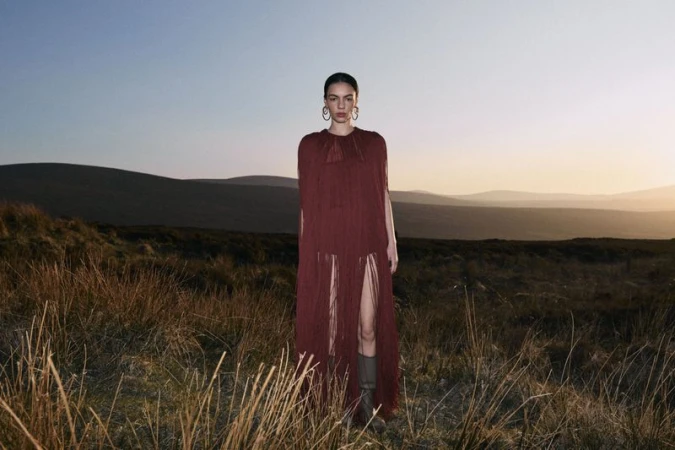 Model wearing a long burgundy fringe dress standing in a grassy field against rolling hills at sunset.