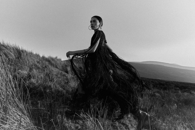 Woman in a black fringe dress walks on a grassy hill in a black and white fashion photograph with a rural landscape background.