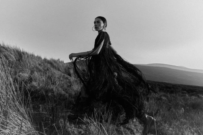 Woman in a black fringe dress walks on a grassy hill in a black and white fashion photograph with a rural landscape background.