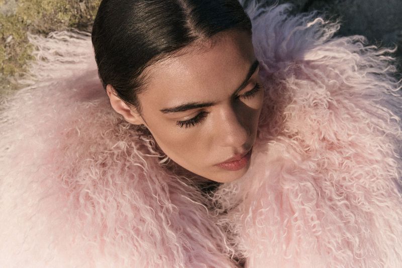 Close-up of a woman with dark hair wearing a light pink shaggy coat, her eyes closed, highlighting a fashion and beauty aesthetic.