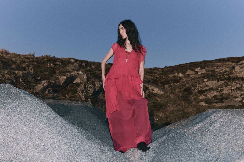 A woman in a red dress poses on a rocky terrain under a twilight sky, creating a striking fashion portrait in a natural setting.