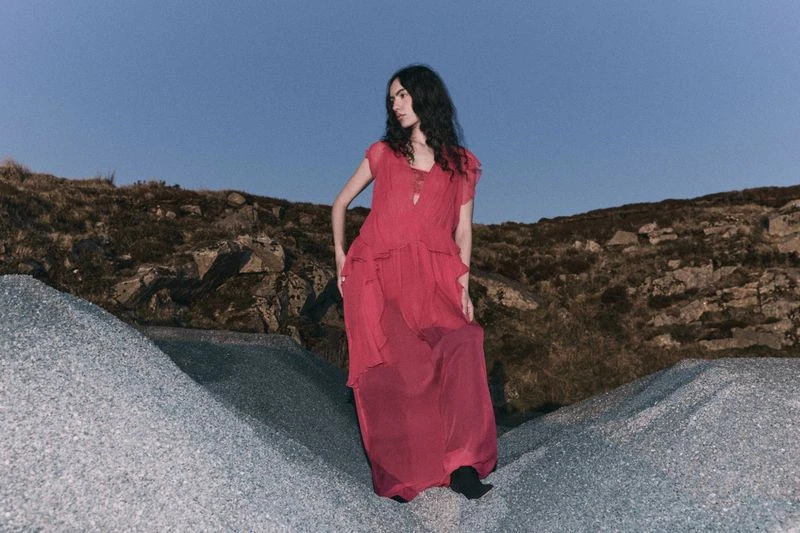 A woman in a red dress poses on a rocky terrain under a twilight sky, creating a striking fashion portrait in a natural setting.