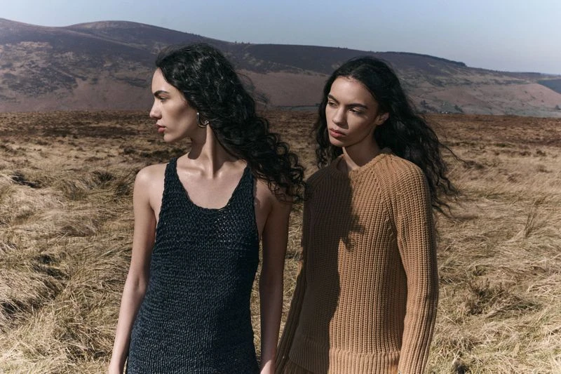 Two women in knitted tops pose outdoors on a grassy plain against a mountain backdrop on a clear day in a medium shot.