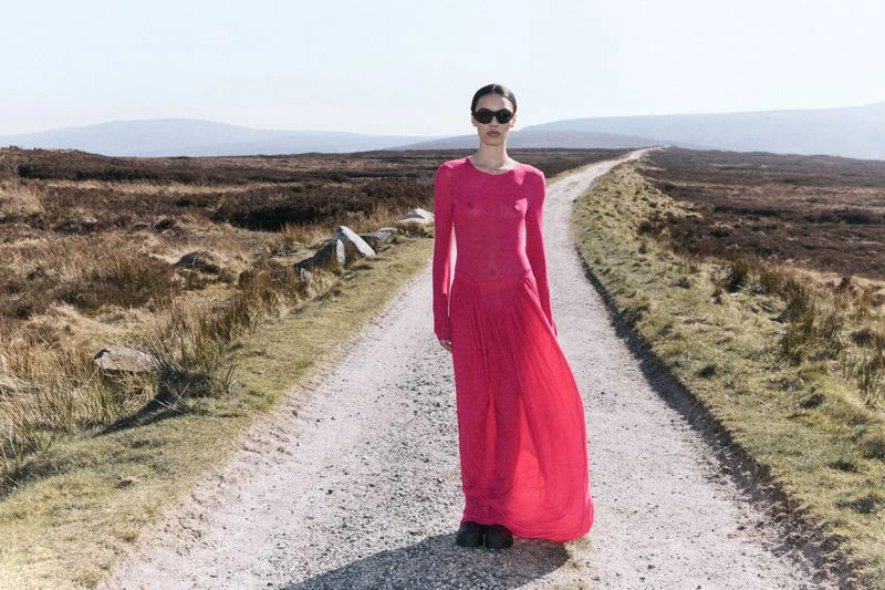 Woman in a long sleeve, floor length magenta dress and sunglasses stands on a path through a field on a sunny day.