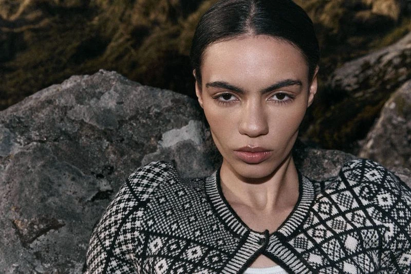 Close-up portrait of a woman with a neutral expression wearing a patterned black and white cardigan in a rocky, outdoor setting.