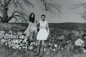 Black and white editorial image of two female models posing outdoors on a weathered stone wall in a rural landscape.