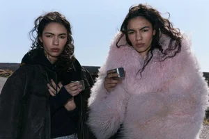 Two women with dark curly hair outdoors, one wearing a pink fuzzy jacket and the other a dark leather jacket holding small metallic cups
