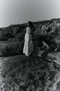Model standing on a large rock in a rural landscape wearing a pale textured top and flowing white skirt.