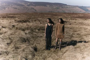 Two female models posing outdoors in a sparse, hilly moorland landscape wearing knitted apparel and tall boots.