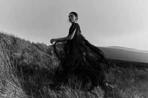 Model in a black fringed dress walking across a grassy hill under an overcast sky for Schön Magazine