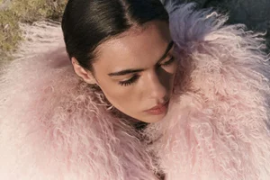 Close-up portrait of a model wearing a fluffy pink textured coat outdoors against natural ground.