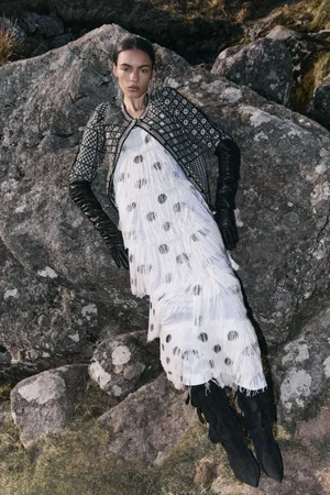Model wearing a patterned jacket, white fringed dress, and black leather gloves reclining against large rocks.